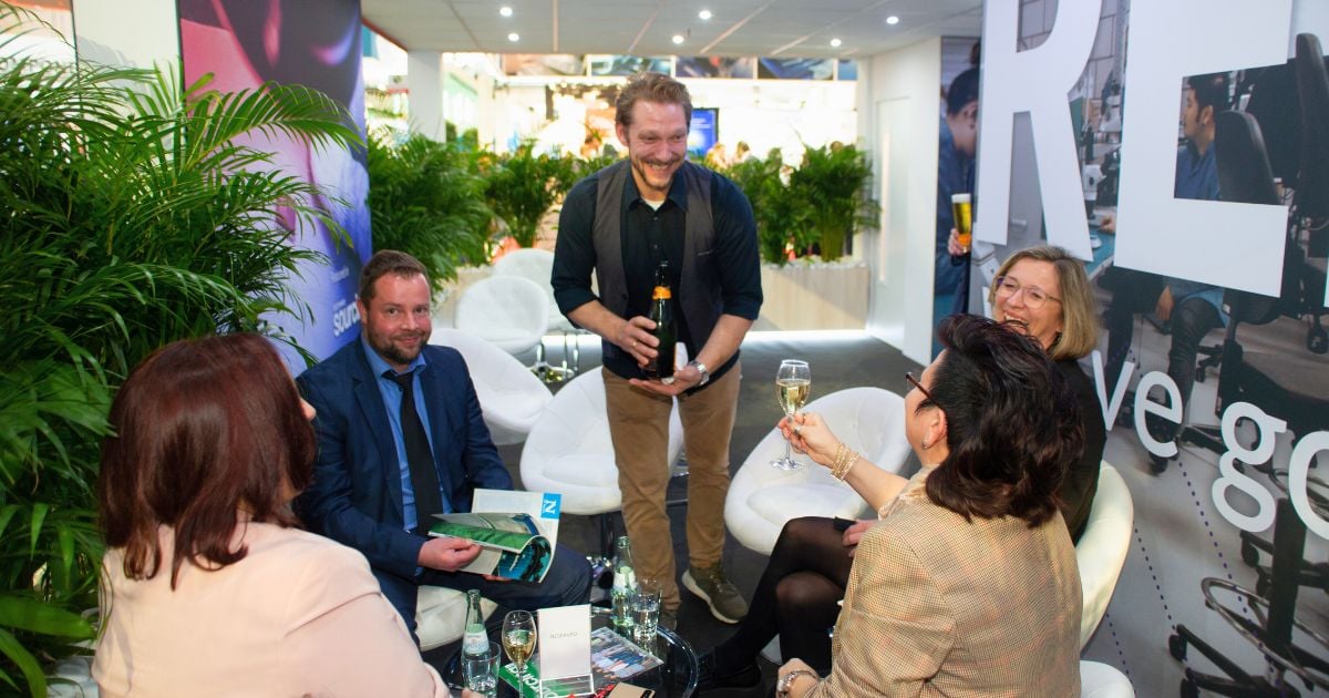 Amidst the hustle and bustle of the electronica show, attendees are seated in a lush, greenery-adorned lounge area of the Velocity Electronics booth. A group of professionals, holding glasses of sparkling wine, engage in cheerful conversation. One man stands, smiling broadly, holding a bottle, adding to the convivial atmosphere. 