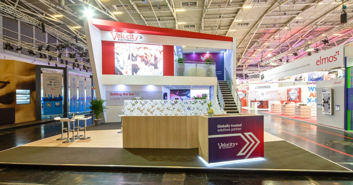 A spacious, well-lit trade show booth for Velocity Electronics, featuring a two-story structure with branding banners, a reception area with white countertops adorned with bird silhouettes, and a presentation screen. The booth is designed to promote Velocity Electronics as a globally trusted solutions partner, emphasizing their role in setting the bar for supply chain solutions.