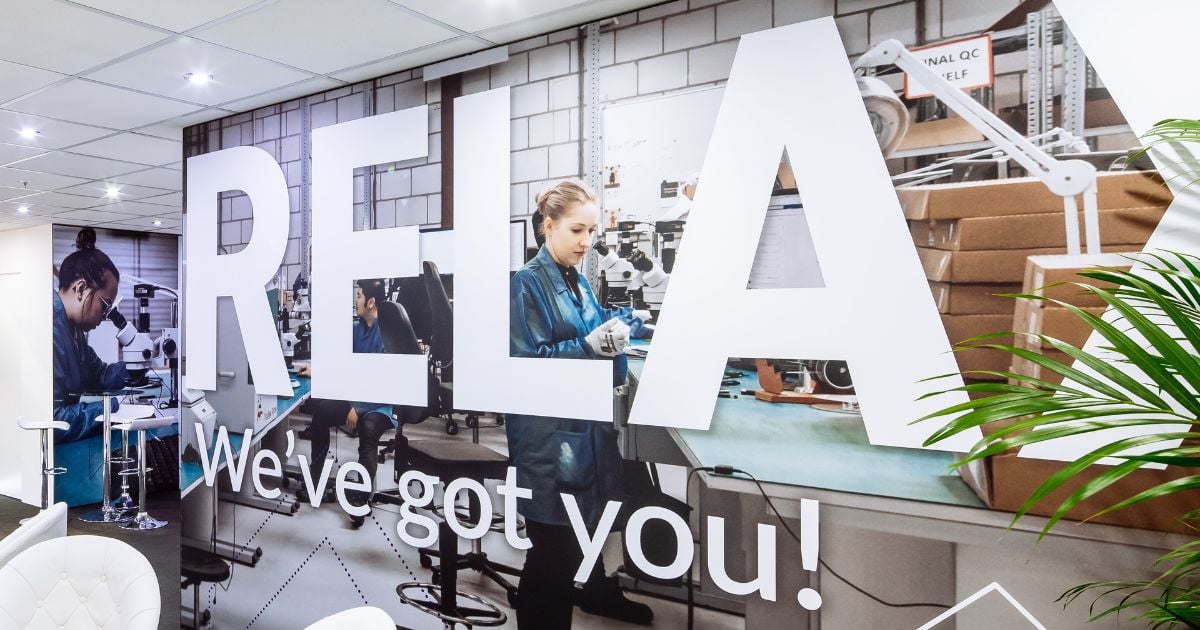 A vibrant wall mural inside Velocity Electronics, featuring large, white letters spelling 'RELAX'. The mural showcases images of focused employees working on electronic components, underlined by the reassuring slogan 'We've got you!'. 