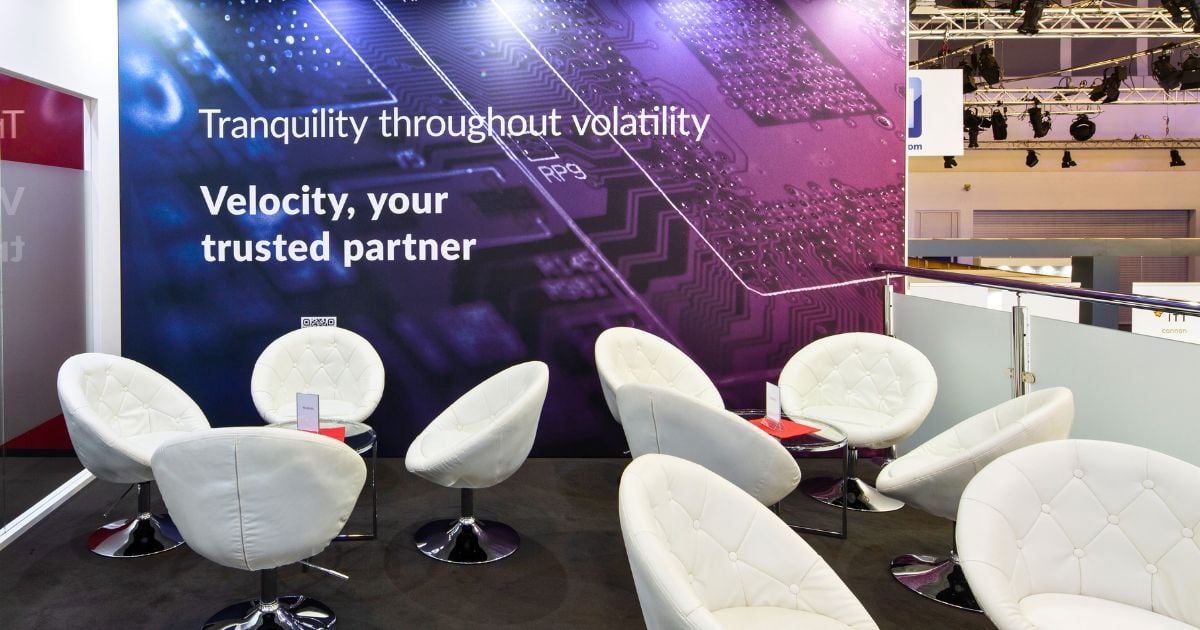 A sleek, modern lounge area within the Velocity Electronics booth at electronica, featuring a backdrop with the message 'Tranquility throughout volatility - Velocity, your trusted partner'. The space is furnished with stylish white egg chairs arranged around small, round tables, creating an inviting setting for discussions. The booth's design emphasizes comfort and the company's commitment to stability in the face of industry challenges.